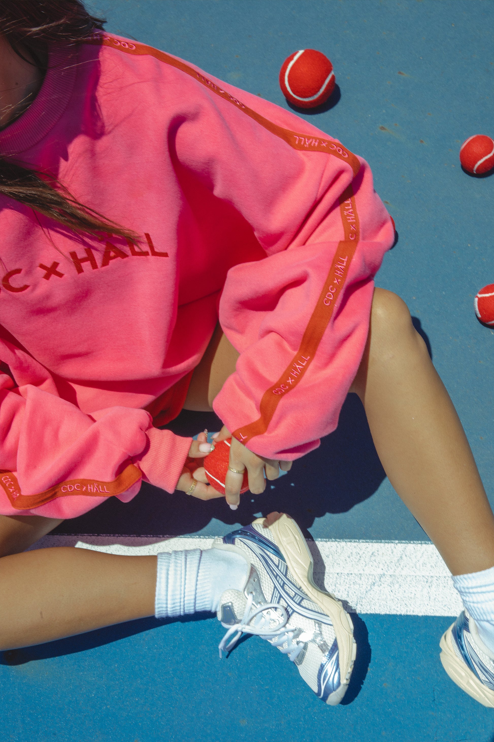 Person wearing a pink sweatshirt with cracker day club x hall text, sitting on a tennis court with tennis balls around.