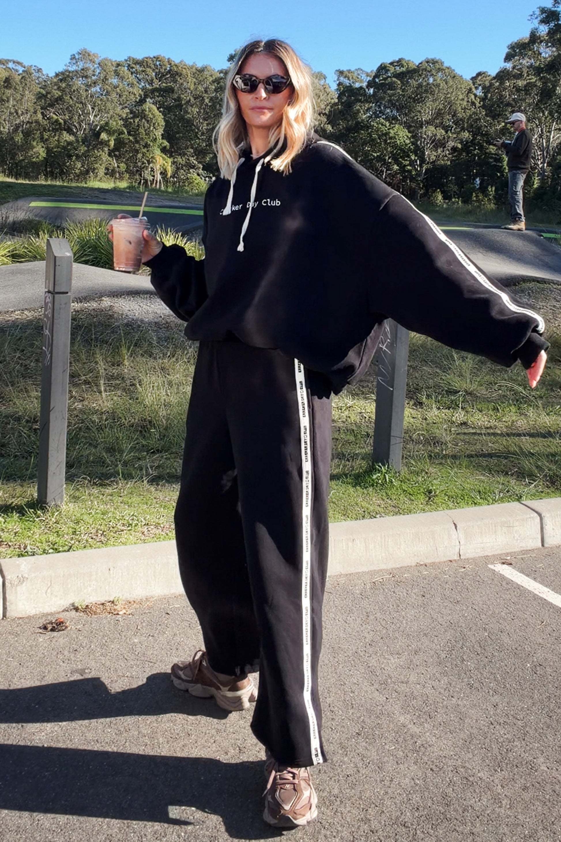 A woman wearing a black oversized hoodie and matching black sweatpants with white side stripes, standing outdoors on a paved area near grass and trees.