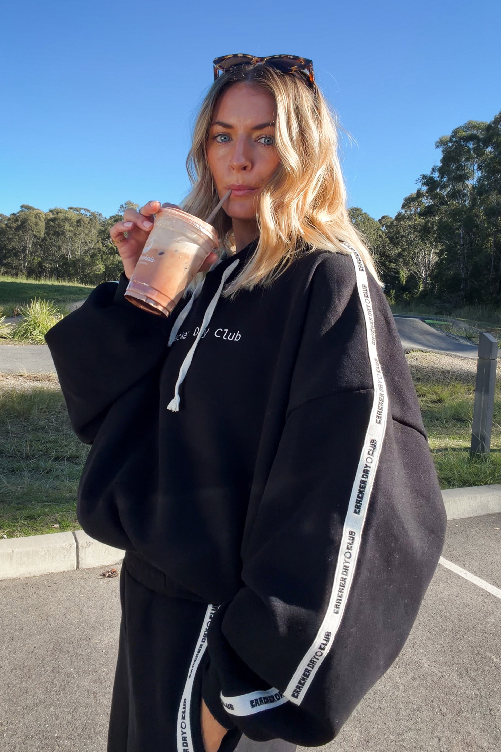 A woman outdoors wearing a black oversized hoodie with white drawstrings and white branded side stripes, holding a drink. The hoodie has white text on the chest reading 'cracker day Club'.