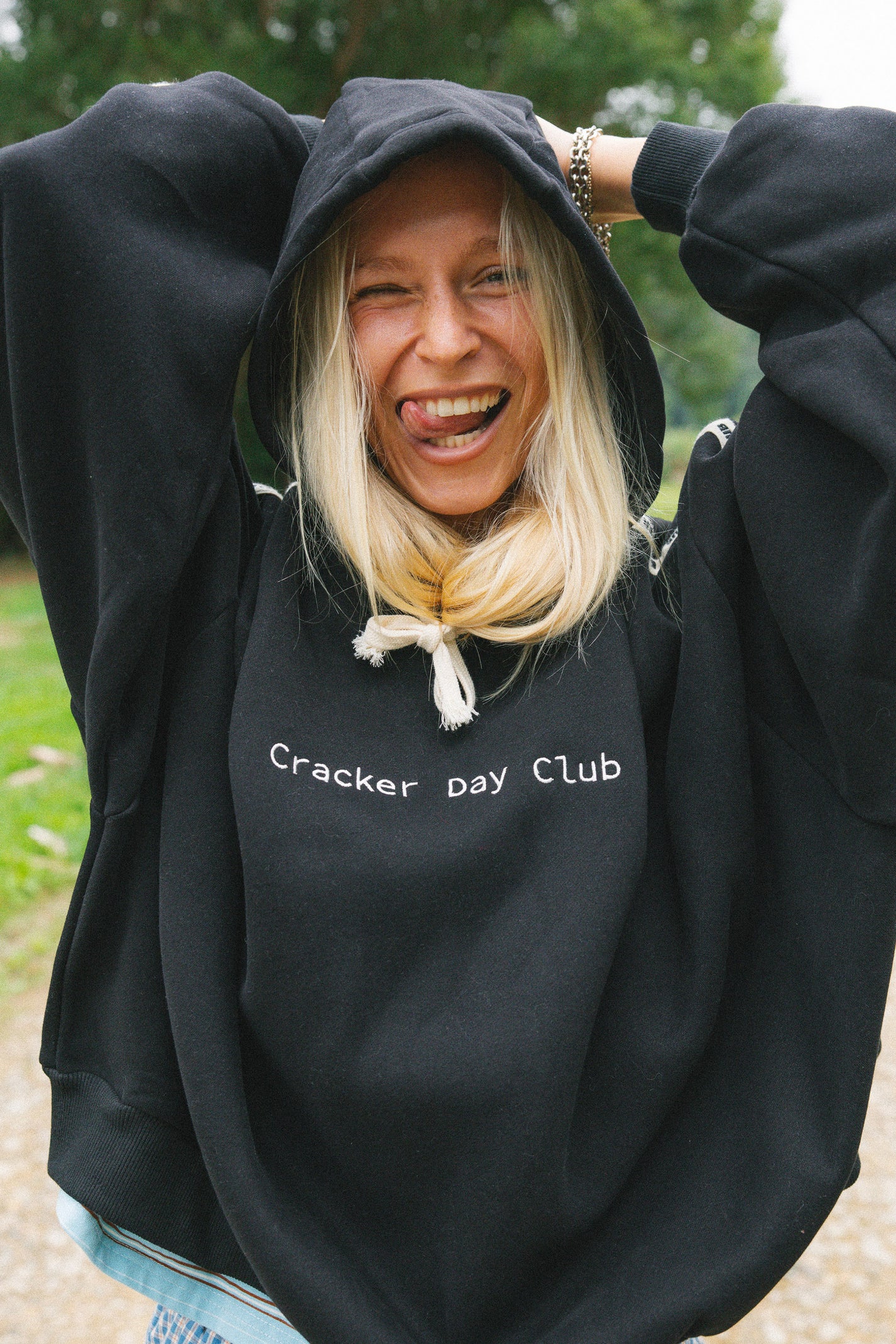 A woman with blonde hair wearing a black hoodie with 'Cracker Day Club' printed in white text on the chest, posing outdoors with her arms raised.