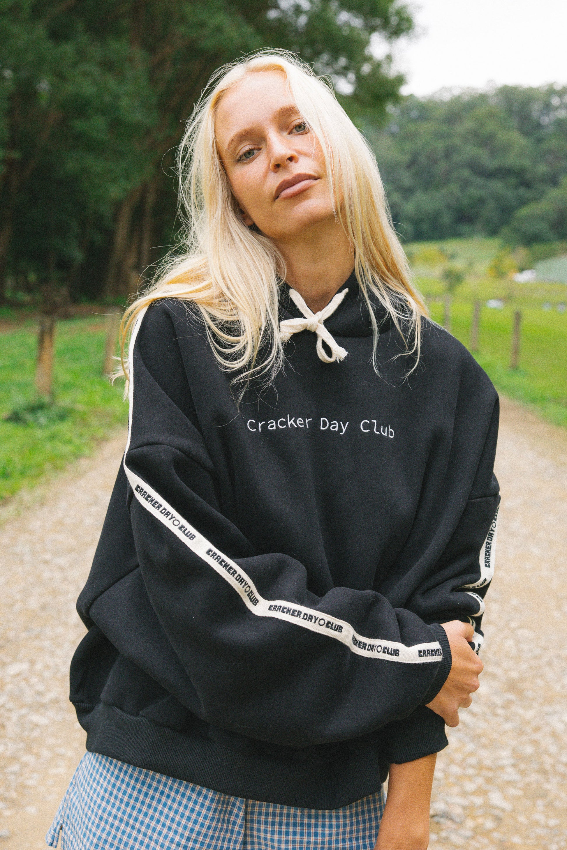 A woman with long blonde hair stands on a gravel path outdoors wearing a black oversized hoodie with white drawstrings, white text reading 'cracker Day Club' on the chest, and white stripe detailing on the sleeves. She is also wearing blue and white gingham pants.
