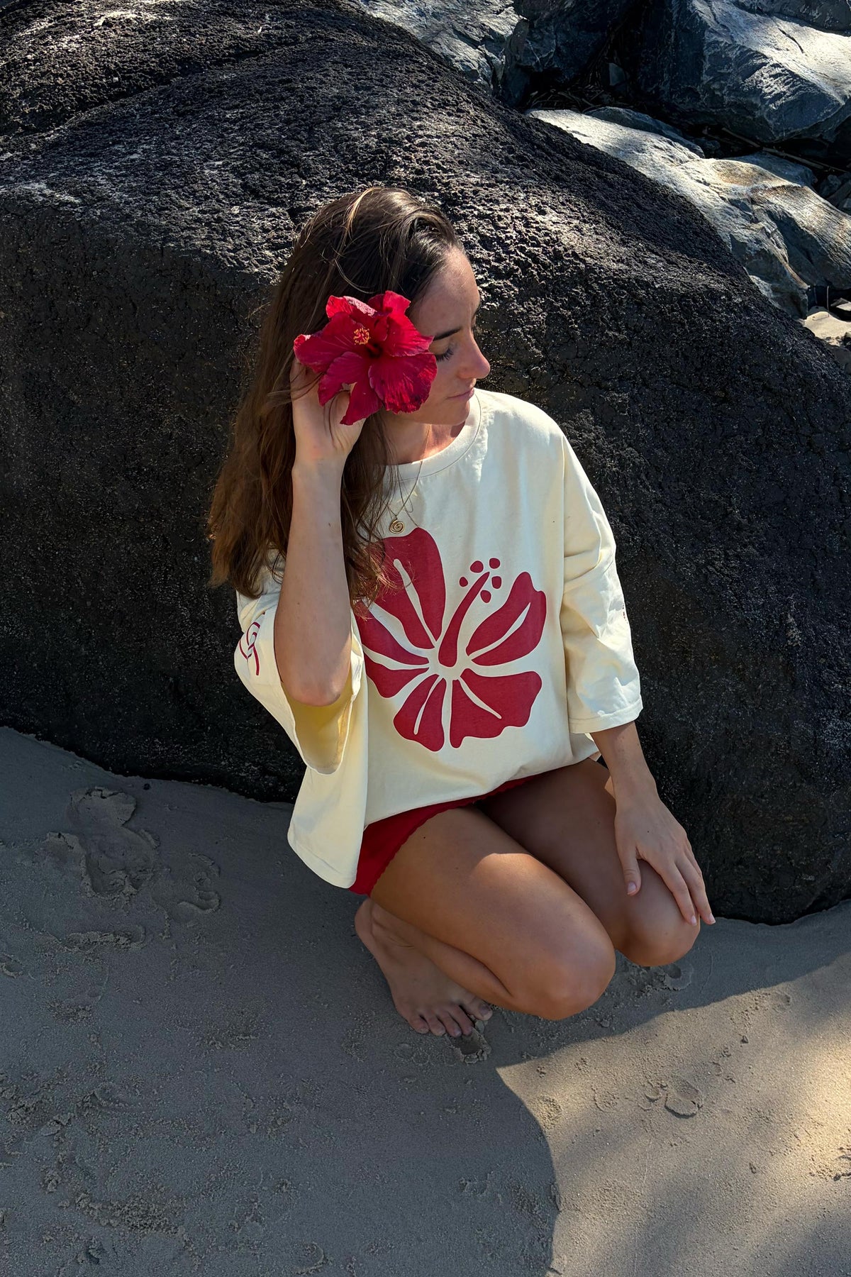 A woman crouches on a sandy beach in front of large dark boulders, wearing a cream short-sleeve top with a large red hibiscus flower graphic and red shorts, while holding a red flower to her eye.