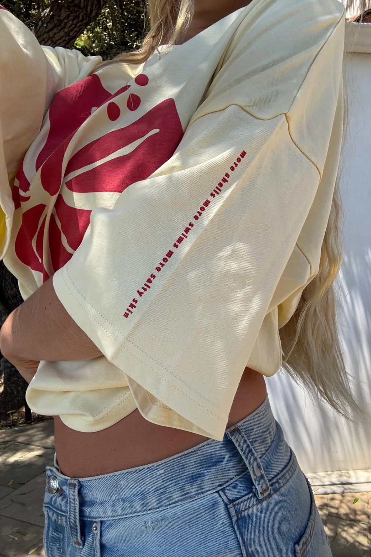 A close-up lifestyle shot of a person wearing a cream-colored oversized graphic t-shirt with a red floral print and text on the sleeve, paired with light blue denim jeans.