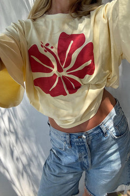 A person wearing a pale yellow cropped T-shirt with a large red hibiscus flower graphic on the front, paired with light blue denim shorts, photographed outdoors against a white wall with soft shadows.