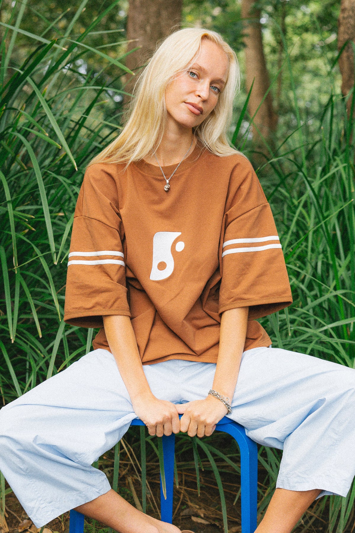 A woman with blonde hair sits on a blue stool in a grassy outdoor setting, wearing an oversized brown t-shirt with a white graphic and white stripes on the sleeves, paired with light blue cropped pants.