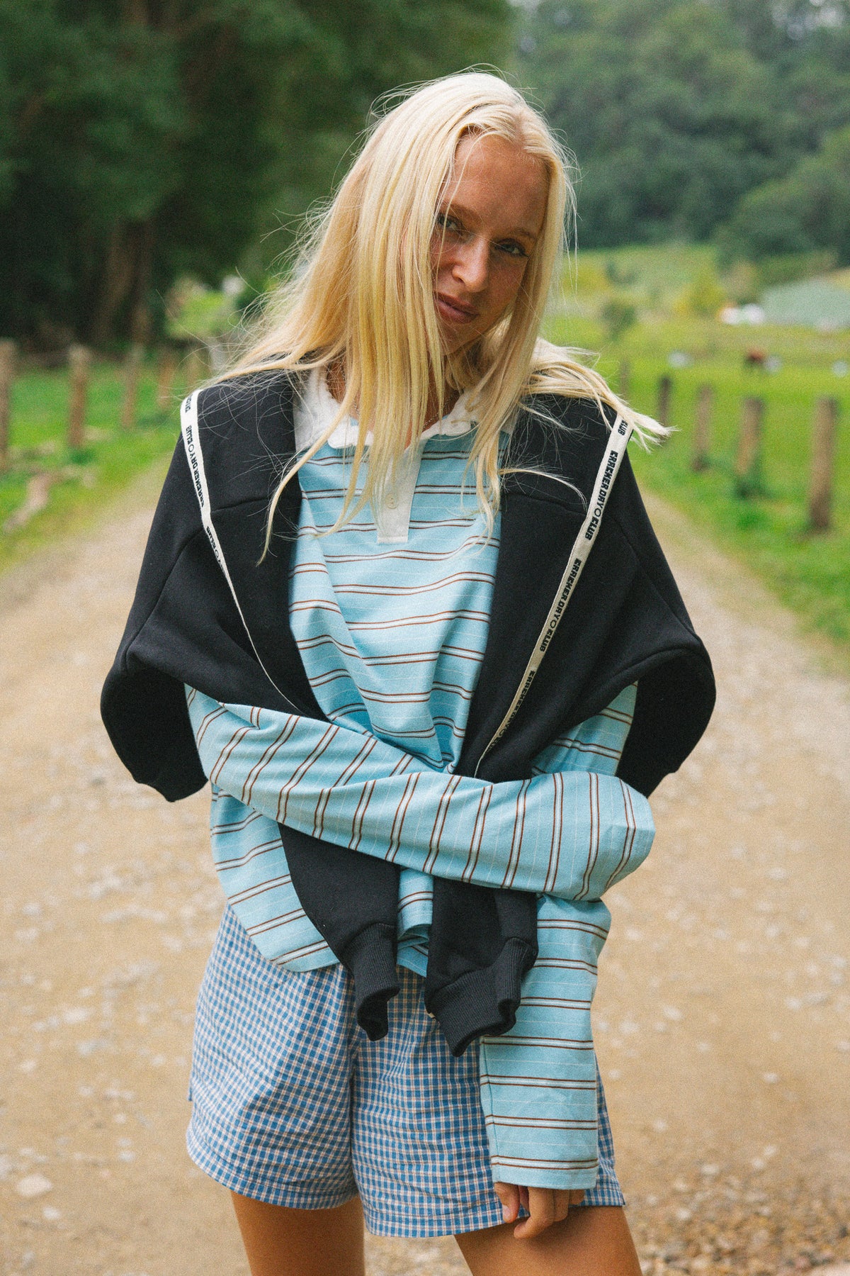 A young woman stands on a dirt path outdoors wearing a light blue and white striped long-sleeve polo top, matching gingham shorts, and a black zip-up hoodie draped over her shoulders.