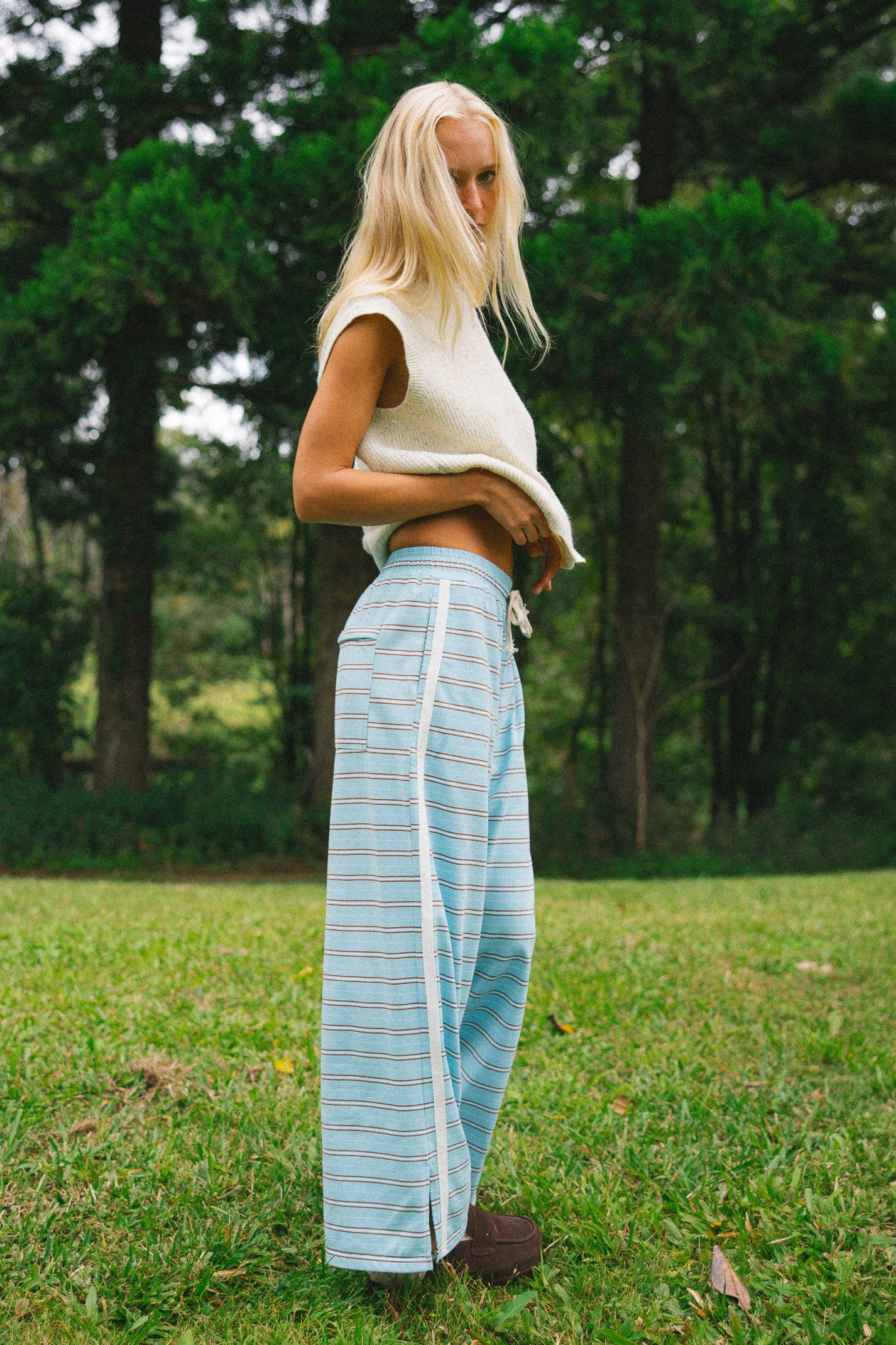 A blonde woman stands on a grassy lawn in front of a forest, wearing a cream sleeveless knit top and light blue wide-leg striped pants.