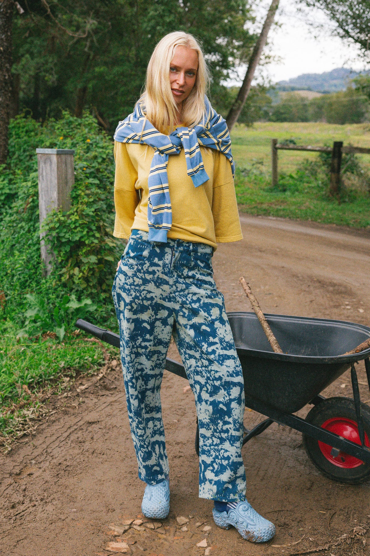 A woman stands outdoors on a dirt path next to a wheelbarrow, wearing a yellow long-sleeve top, blue-and-white patterned jeans, a blue-and-white striped knit scarf, and blue knit shoes.