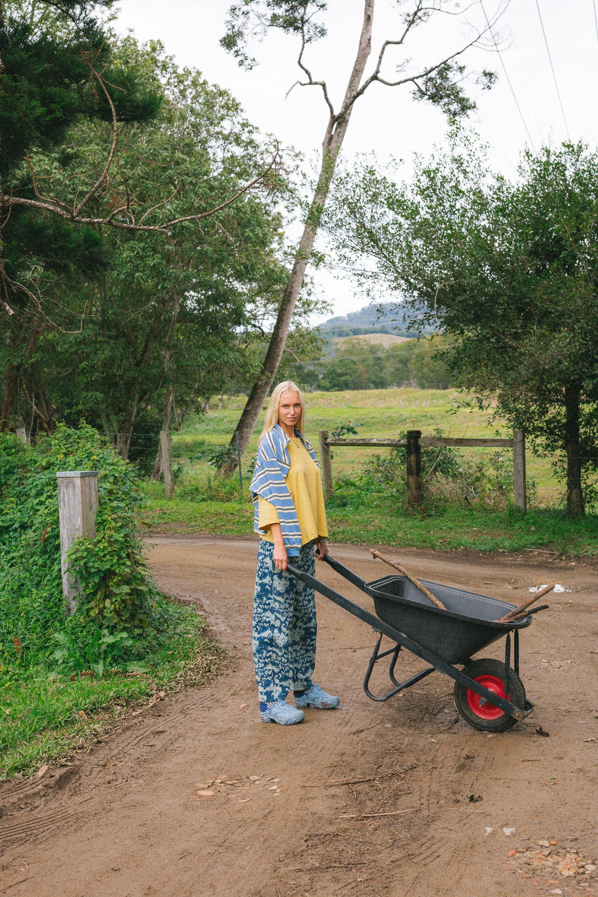 A woman stands on a dirt path outdoors, holding the handle of a black wheelbarrow with a red wheel. She is wearing a blue and white striped shirt, a yellow vest, blue patterned pants, and blue work boots.