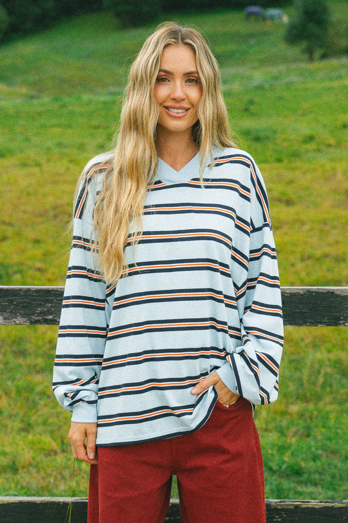 A woman with long blonde hair stands outdoors in front of a wooden fence and green grass, wearing a light blue long-sleeve striped shirt with navy, black, and tan stripes, paired with rust red pants.