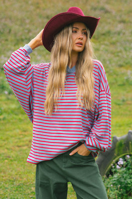 A woman wearing a red and blue striped long-sleeve shirt, dark green pants, and a burgundy wide-brim hat, standing outdoors in a grassy field.