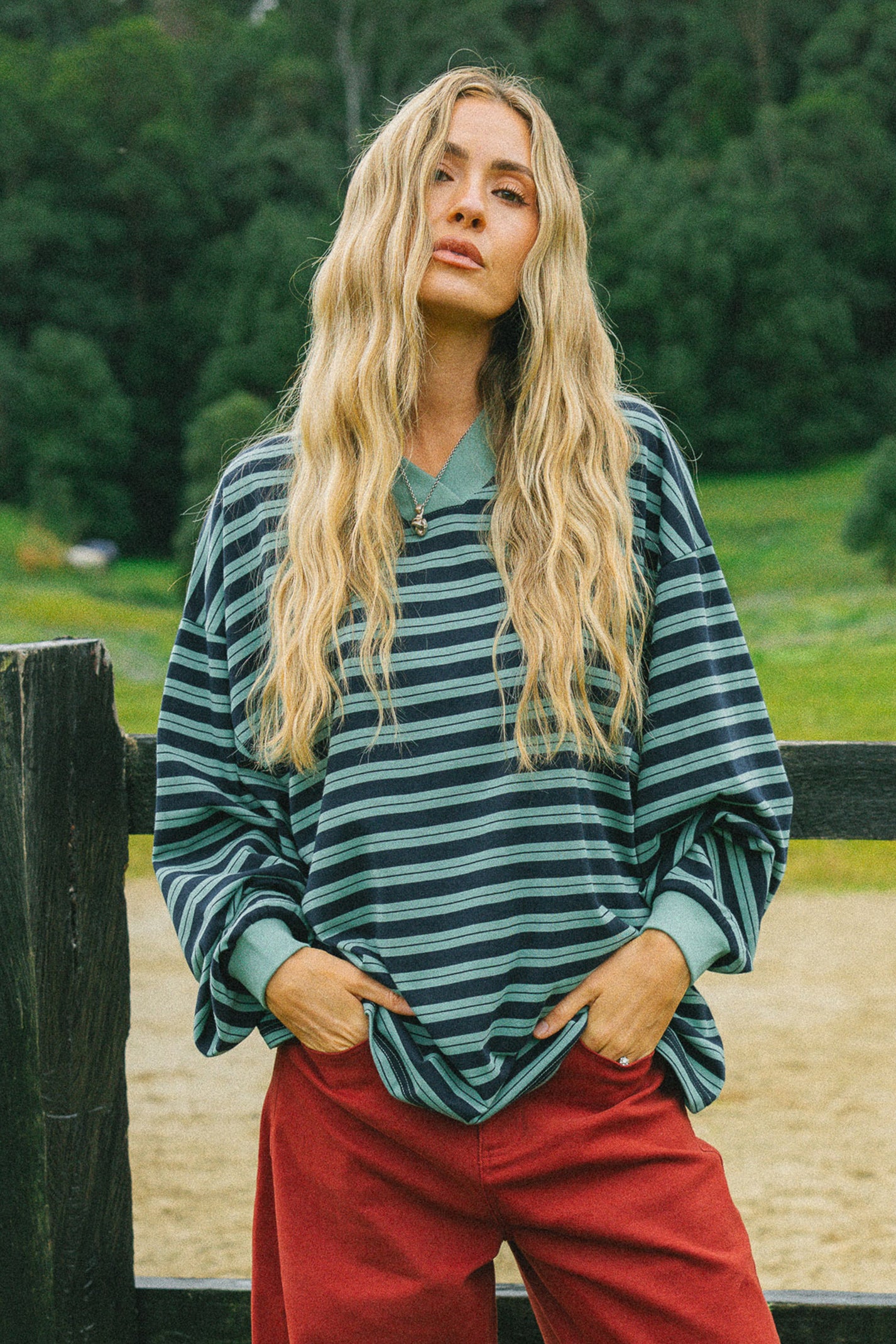 A woman with long blonde hair stands outdoors in front of a wooden fence, wearing a loose long-sleeve striped top in dark teal and green with red pants.