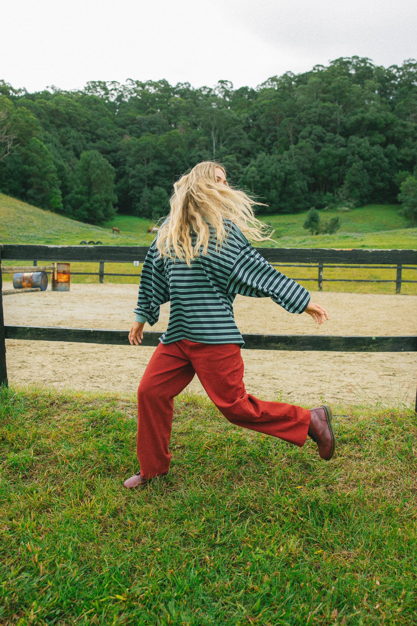 A woman with long blonde hair runs across a grassy field, seen from behind. She is wearing a dark green and white striped long-sleeve top, rust-red pants, and brown shoes, with a fence, sandy arena, and forest in the background.