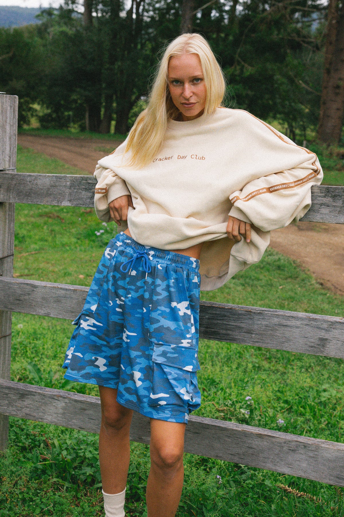 A woman stands outdoors in front of a wooden fence wearing an oversized cream sweatshirt with 'Cracker Day Club' printed on the chest and blue camouflage shorts.