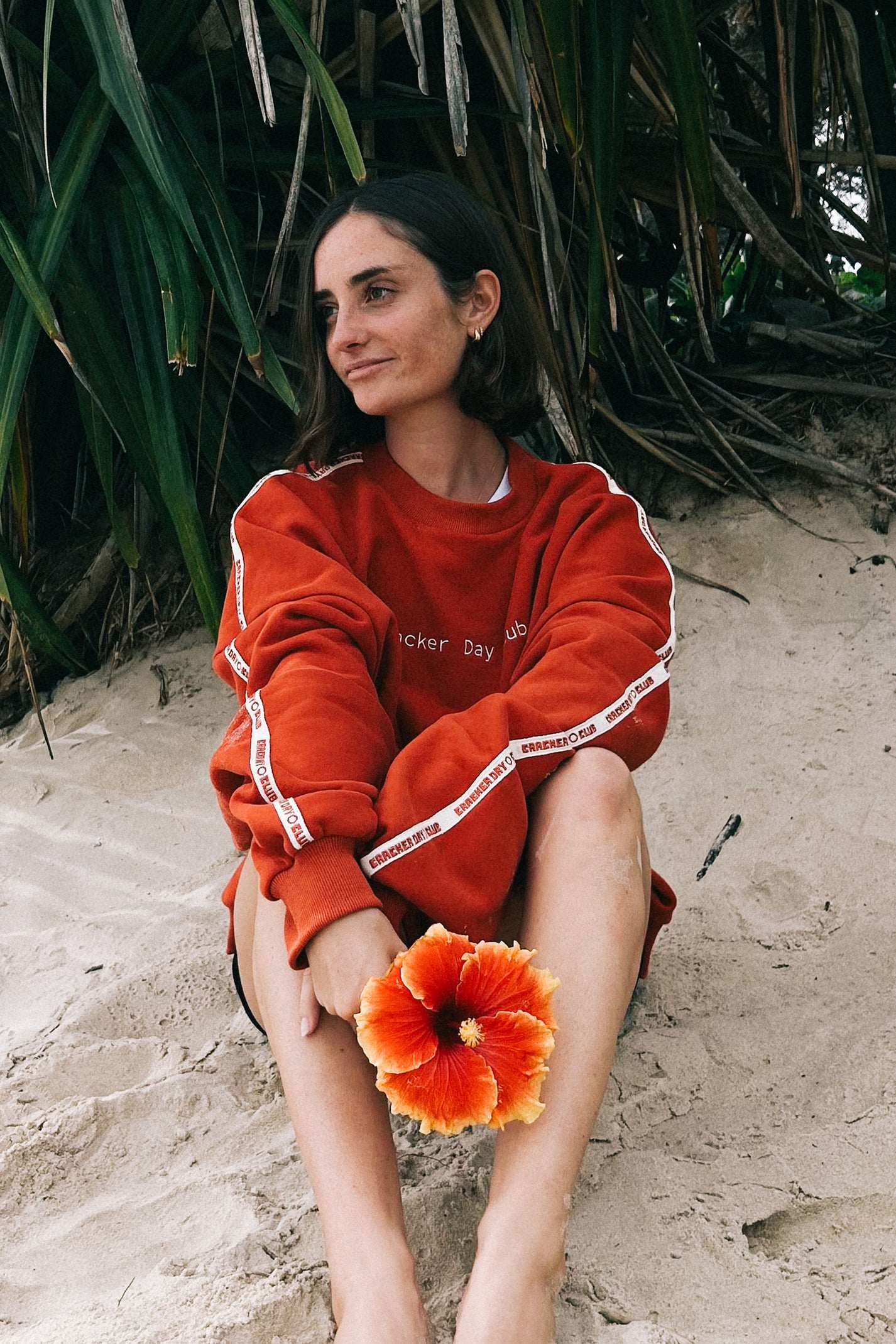 Woman in a cracker day club red sweatshirt sitting on sand with a large orange flower.