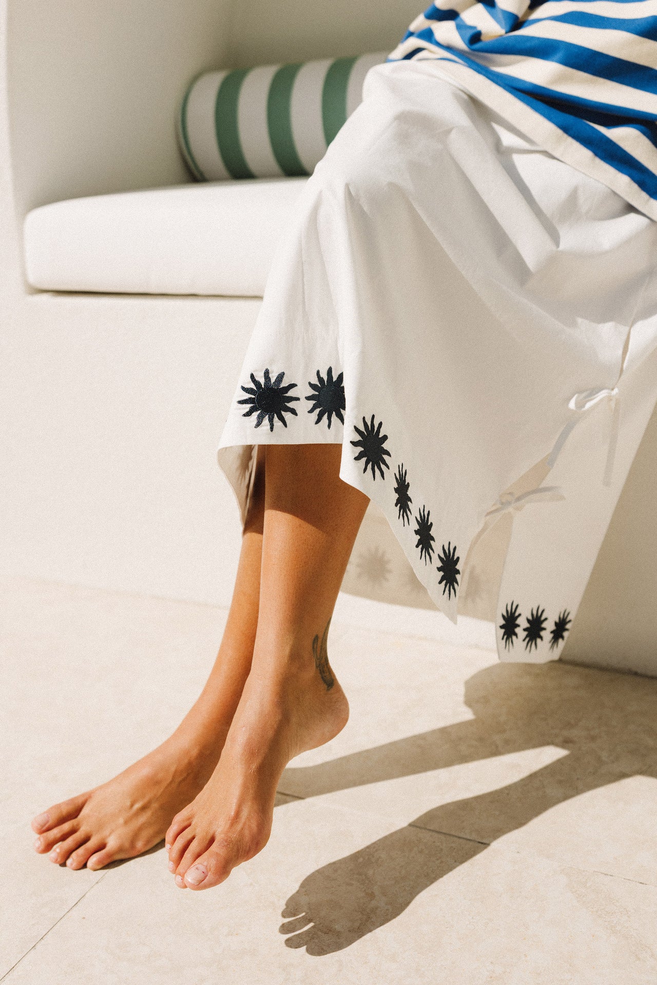 Woman in a cracker day club blue and white striped shirt and white maxi skirt with sun patterns sitting on a white surface.