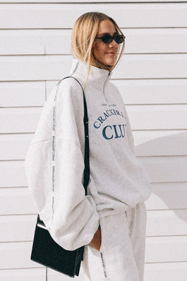 Person wearing a white sweatshirt with text and pants, holding a black bag against a light wooden wall.